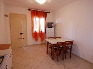 A small, clean room with a tiled floor, a wooden dining table with four chairs, a white refrigerator, and a door with a curtain.