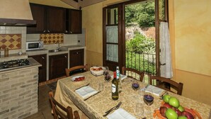 A kitchen with a dining table set for two, a gas stove, a microwave, and a view of the garden through an open door. - Farmhouse in Montoro Near Montoro Castle (Narni)