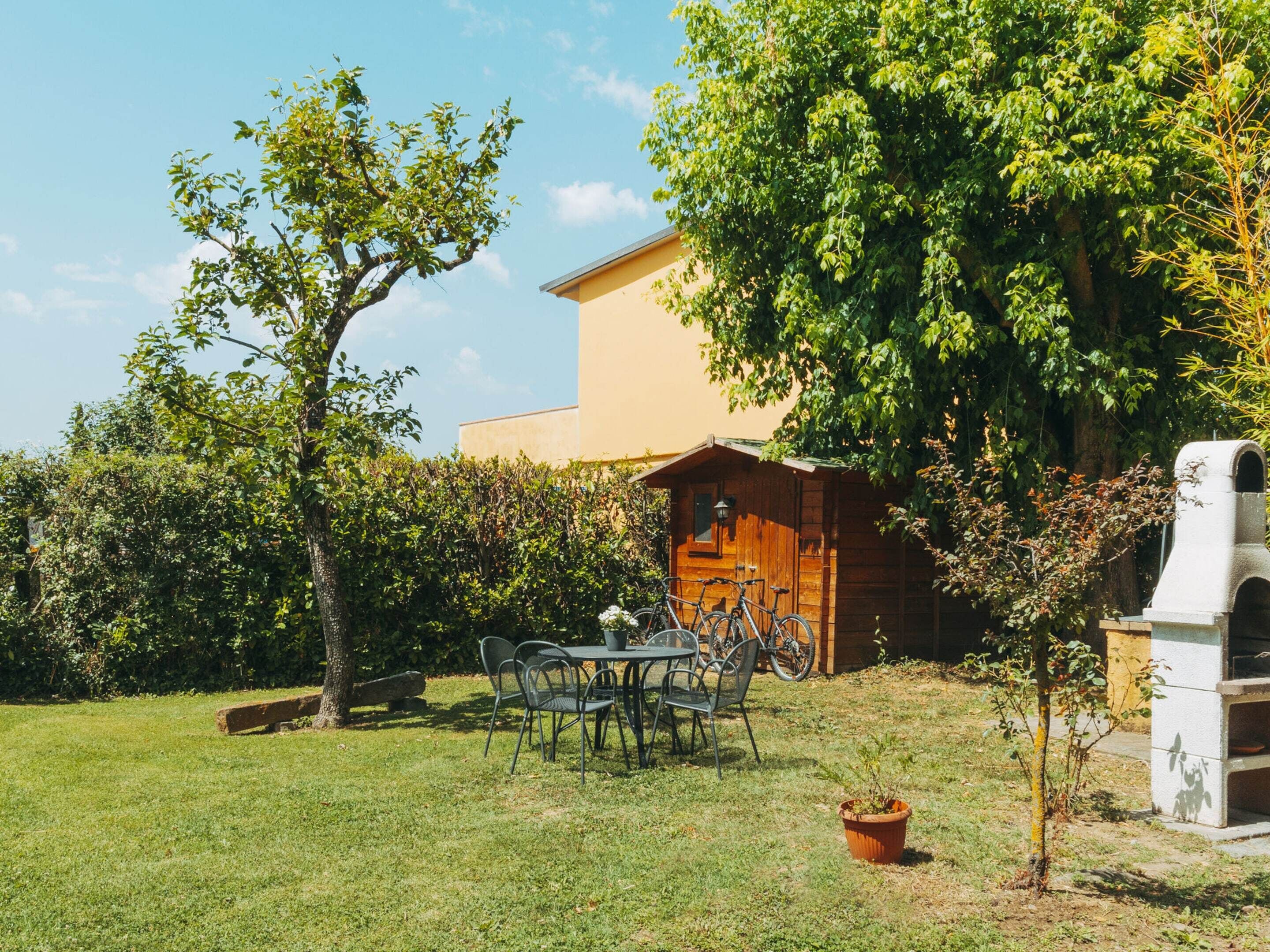 A wooden cabin with a table and chairs in a grassy area, surrounded by trees and a building in the background.