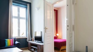 Una habitación con suelo de parqué, un televisor sobre un mueble de madera, una cama con una manta roja y una ventana con cortinas.
