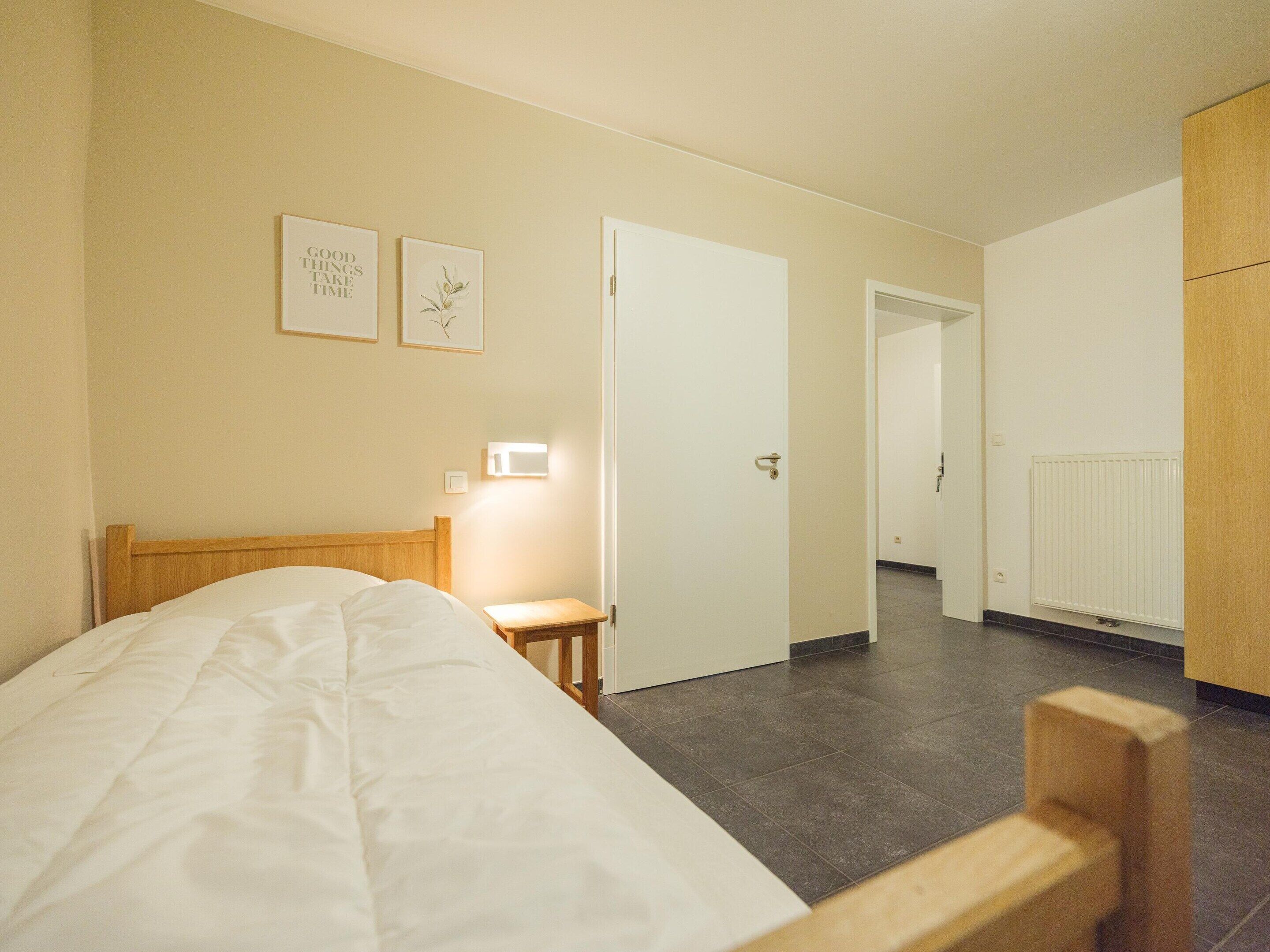 A bedroom with a bed, a bedside table, a nightstand, a wall-mounted light, and a door leading to another room.