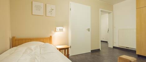 A bedroom with a bed, a bedside table, a nightstand, a wall-mounted light, and a door leading to another room.