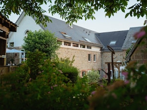 A stone building with a sloped roof, a wooden pergola, and a garden area with bushes and flowers.