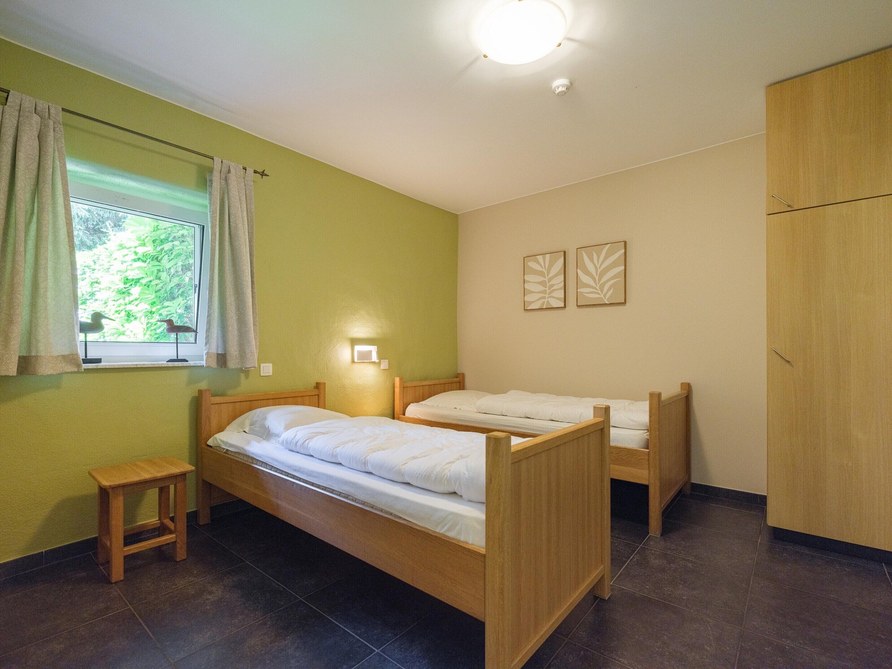 Ein Zimmer mit zwei Holzbett, einem kleinen Hocker, einem Fenster mit Vorhängen, einer Wandleuchte und einem Kleiderschrank.