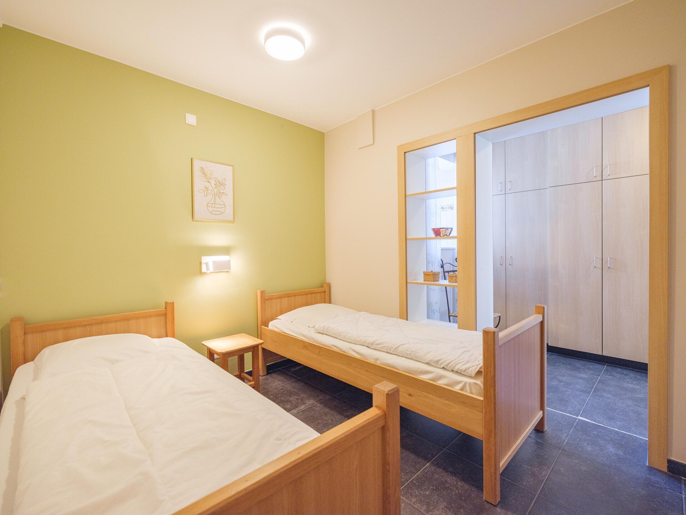 Un dormitorio con dos camas de madera, una mesita de noche pequeña, una lámpara de pared, un estante con cestas y un armario con puertas correderas.