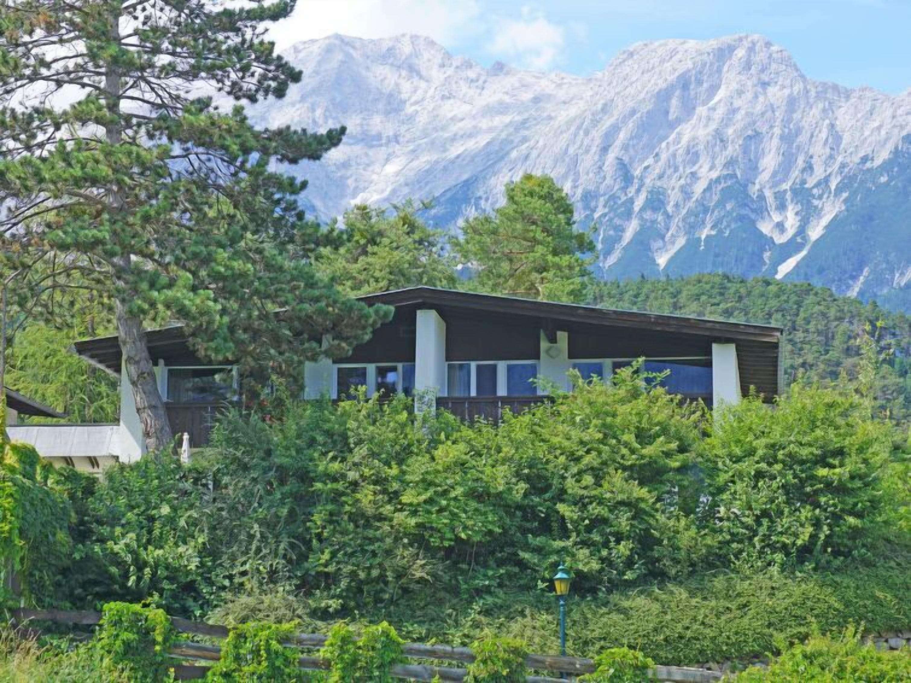 A modern building with a flat roof, surrounded by dense greenery and mountains in the background.