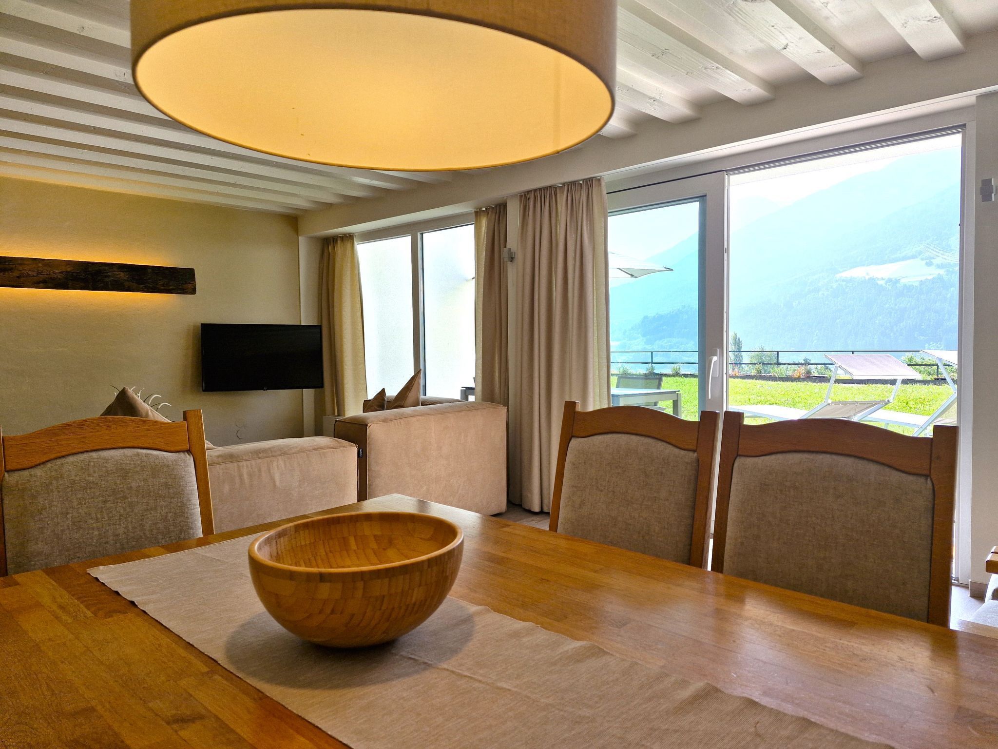 A dining area with a wooden table, chairs, a wooden bowl, a flat-screen TV, and a large window with a view of the outdoors.