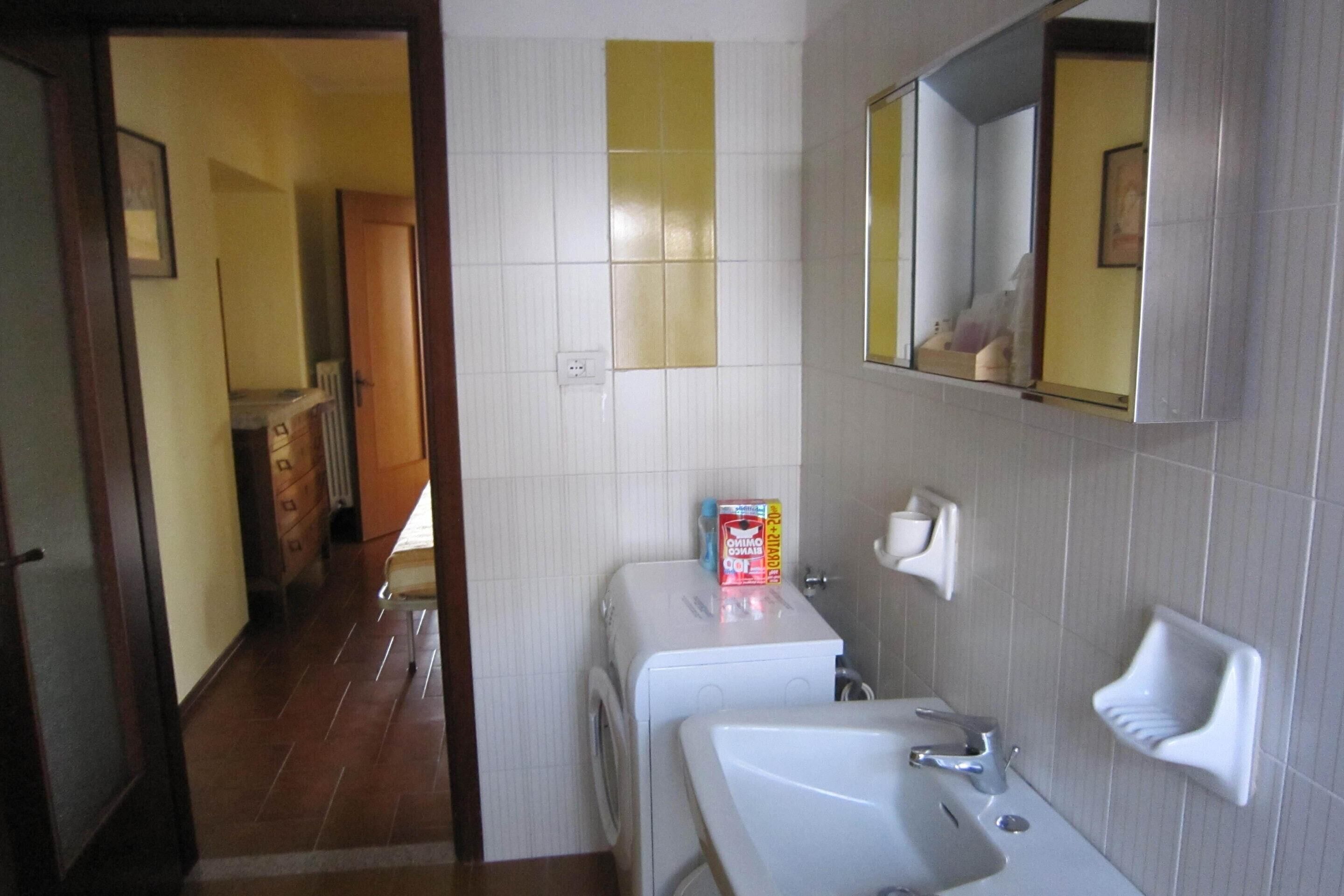 A bathroom with a white sink, mirror, and a towel rack. There is a washing machine and a box of laundry detergent on the counter.