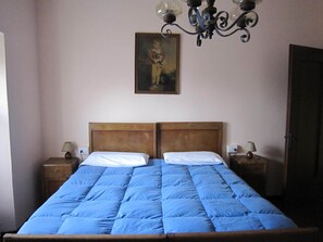 Una cama doble con colcha azul, cabecera de madera, dos mesitas de noche con lámparas, un cuadro colgado en la pared y una araña.