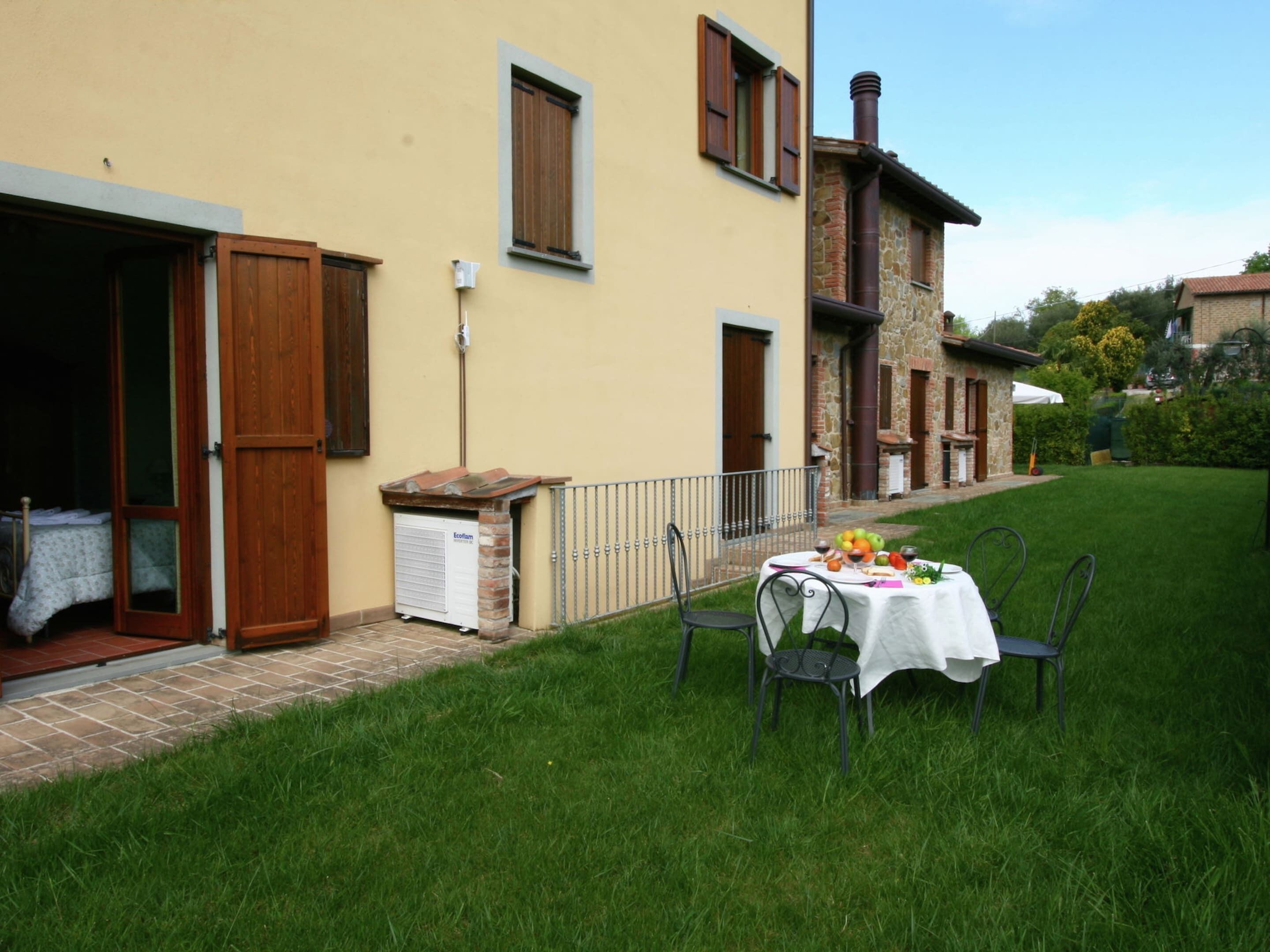 A cozy outdoor dining area with a table set for two, surrounded by a well-maintained lawn and a stone building with wooden shutters.