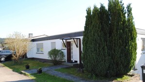 Een gelijkvloers huis met een veranda, een oprit en een hoge naaldboom.