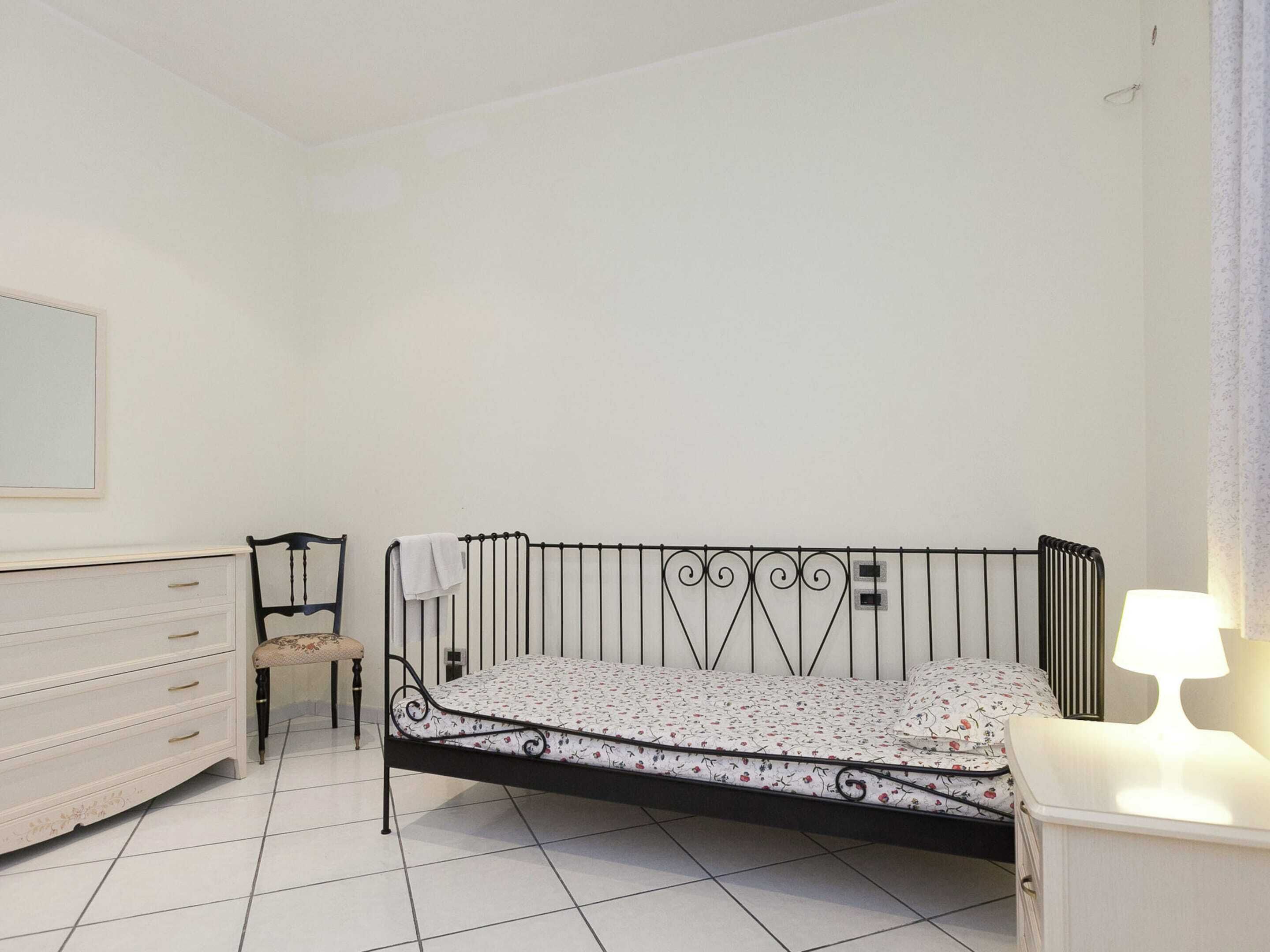 Una camera da letto con una struttura letto in metallo, una cassettiera bianca, una sedia e un comodino con una lampada.