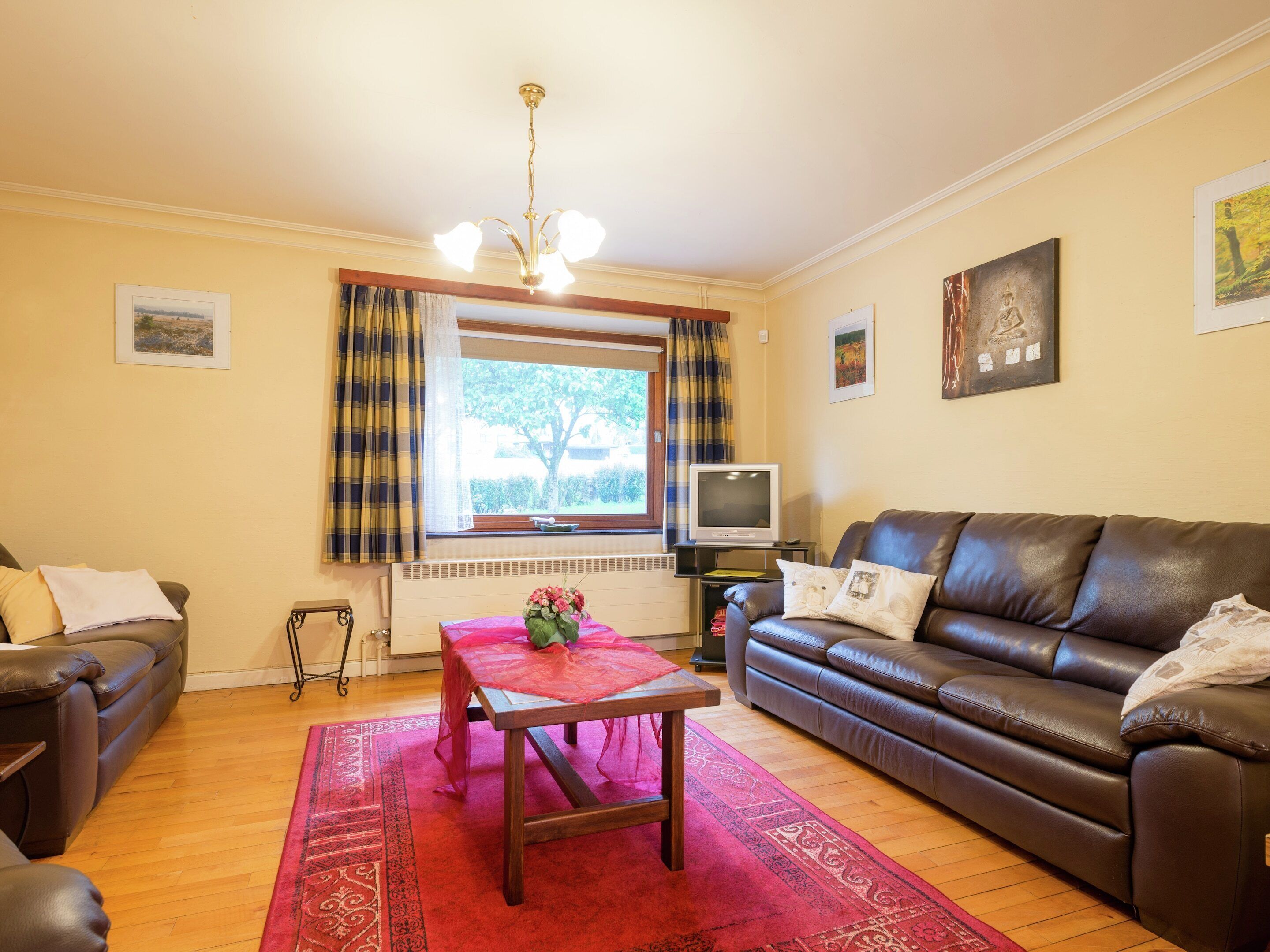 A living room with a brown leather sofa, a wooden coffee table, a red rug, a television on a stand, and a window with curtains.