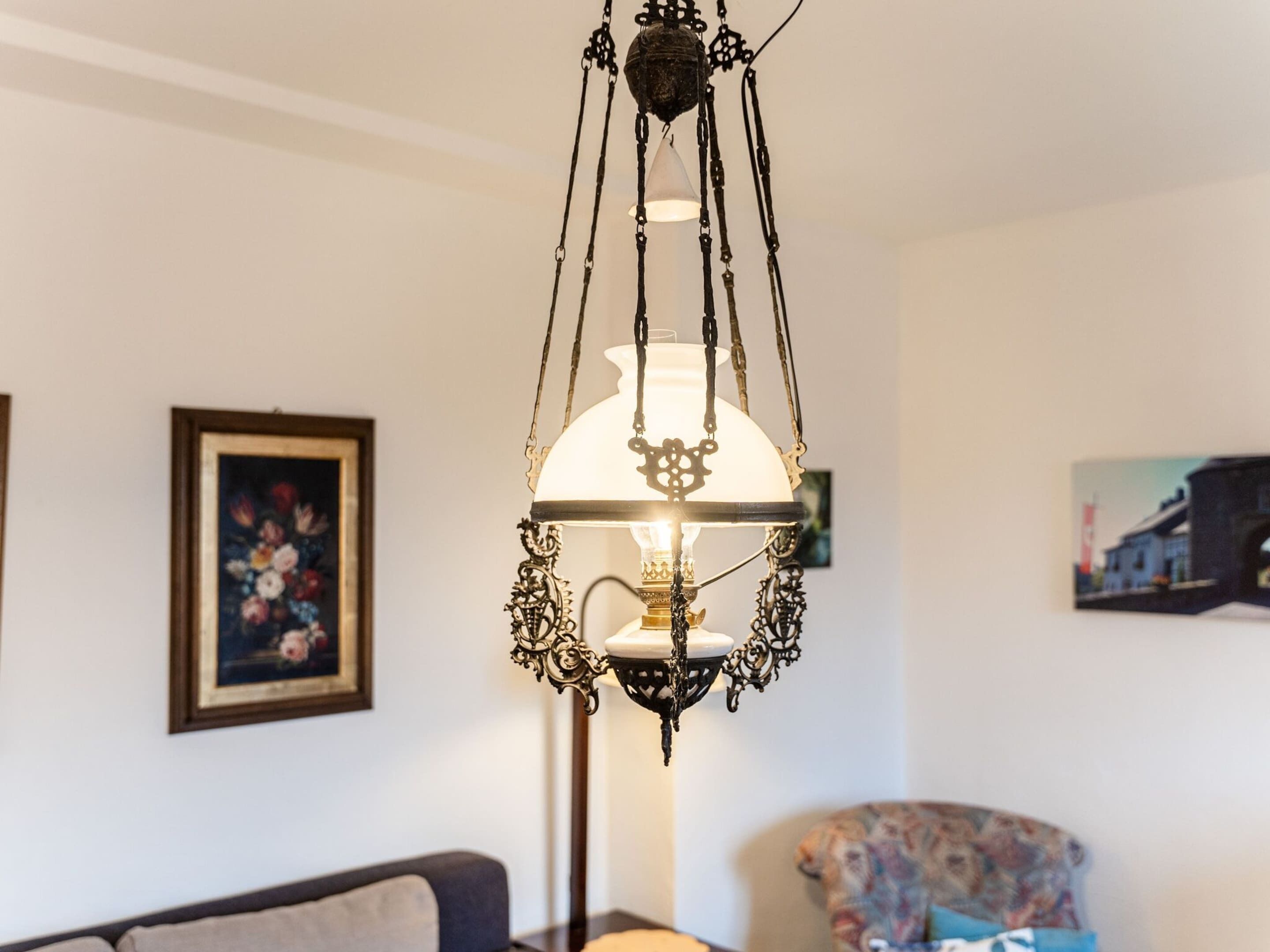 A chandelier with intricate metalwork and a white lampshade, hanging in a room with a sofa and a framed painting on the wall.