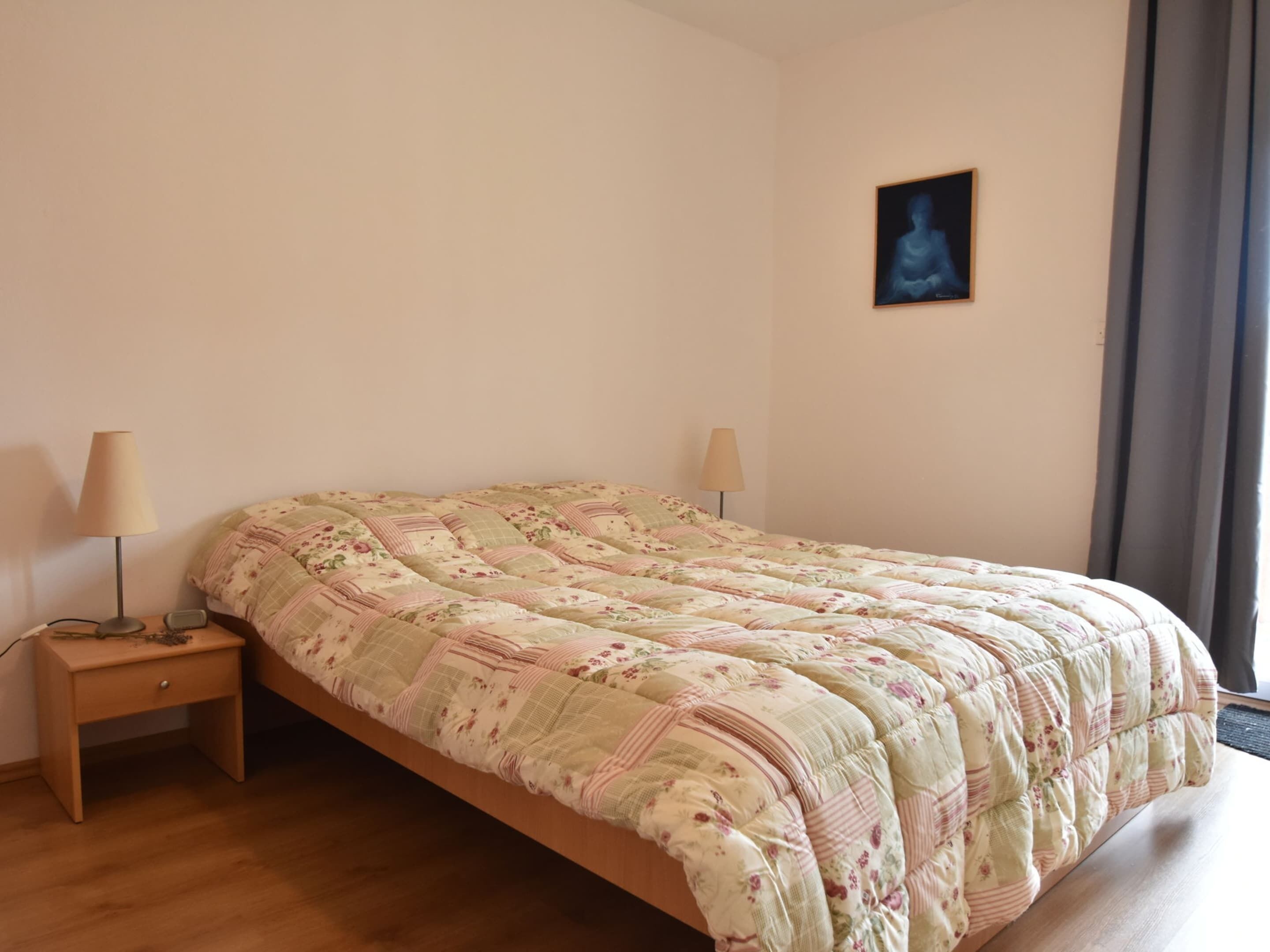 A bedroom with a floral-patterned bedspread, two bedside lamps, a small wooden nightstand, and a framed picture on the wall.