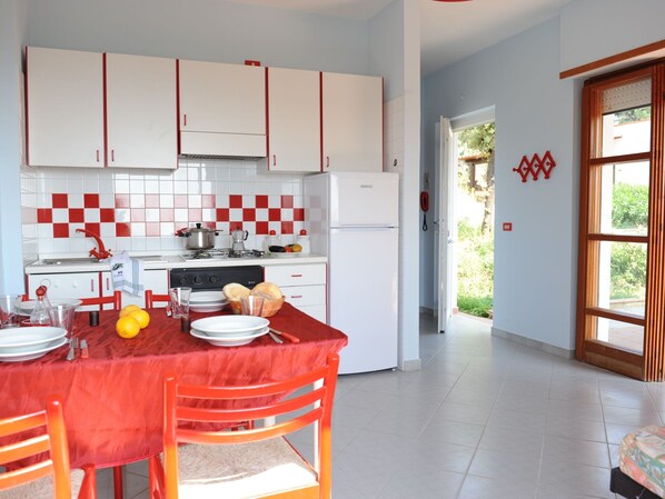 Une cuisine avec des meubles blancs et des touches de rouge, une table à manger avec des chaises, et une vue sur l’extérieur par une porte.
