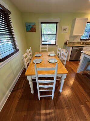 A dining area with a wooden table and white chairs, a window with blinds, and a kitchen with a dishwasher. - Swept Away One -Great Family Beach Home (Kill Devil Hills)