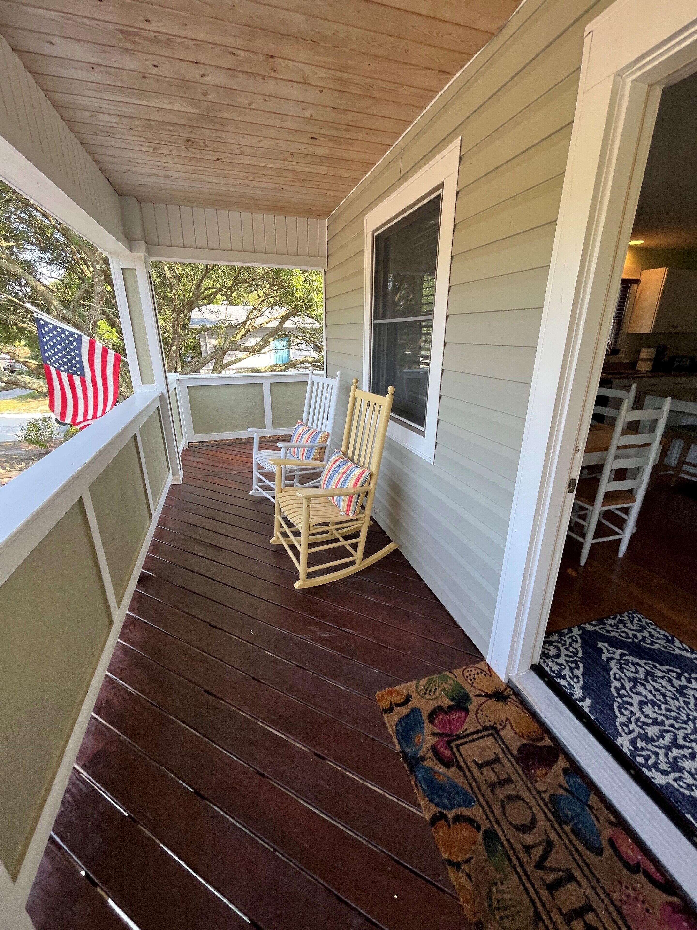 A wooden porch with a rocking chair and a striped cushion, a large window, and a welcome mat.