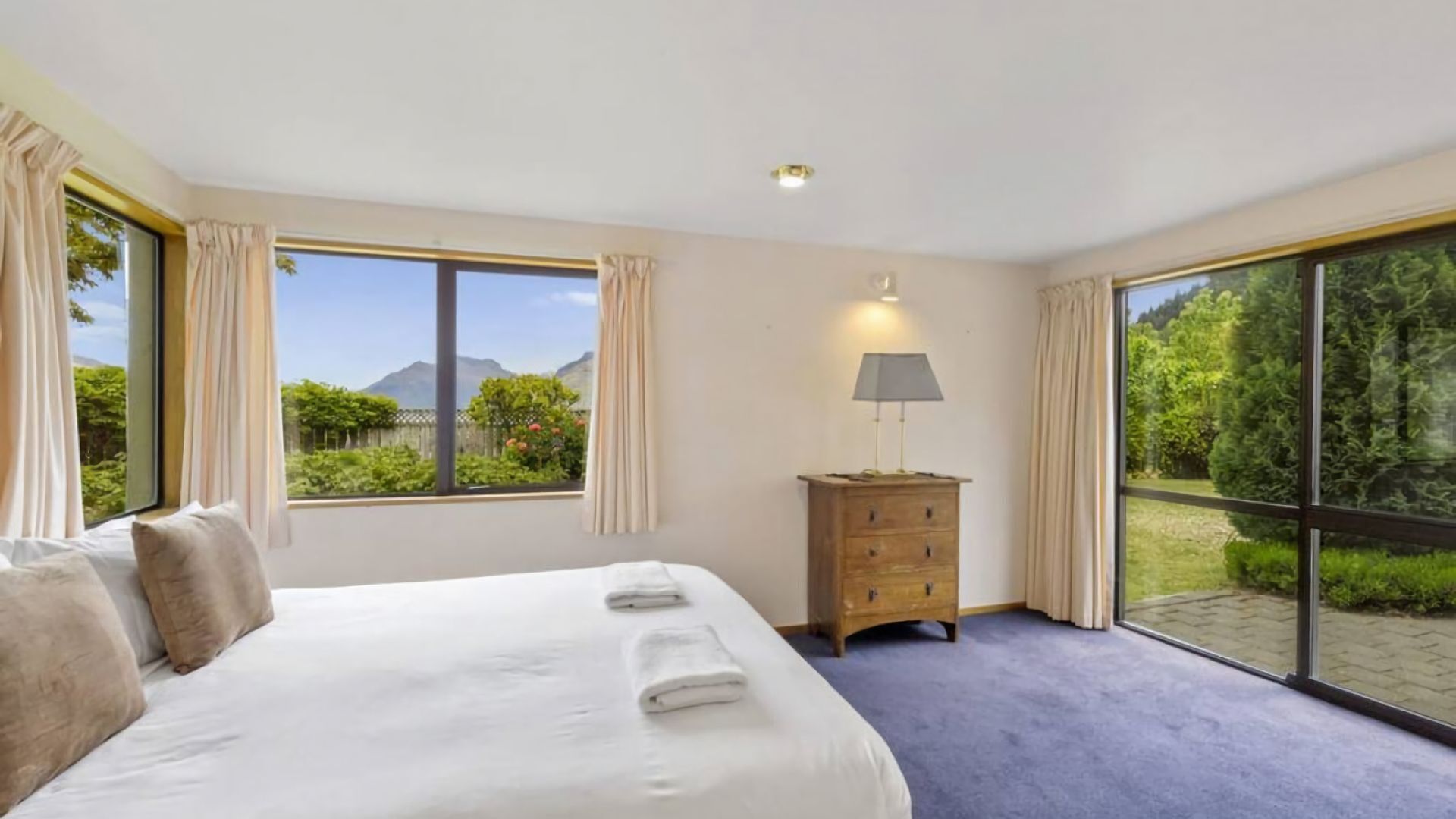 Une chambre avec un grand lit, une commode en bois et une vue sur la verdure et les montagnes par les fenêtres.