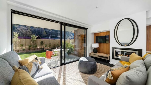 Una sala de estar moderna con un amplio ventanal corredizo que da a un jardín. Hay una chimenea, un televisor y un espacio de descanso cómodo con sofás y una mesa de centro.