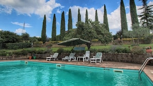 En swimmingpool med solstolar och en pergola. Höga träd och en mur av tegelstenar syns i bakgrunden.