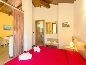 A hotel room with a red bedspread, a wooden ceiling, a desk, a chair, a bathroom with a mirror, and towels on the bed.