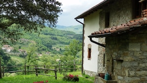 A rustic stone building with a white facade and a red-tiled roof, surrounded by lush greenery and a view of a valley with scattered houses. - Charming Tuscan Farmhouse (Ortignano Raggiolo)