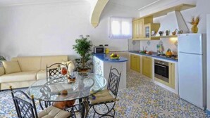 A compact living space with a round glass table, a sofa, and a kitchen area with a refrigerator, stove, and cabinets. - Apartment near Praia with Large Terrace & Spectacular Views (Praiano)