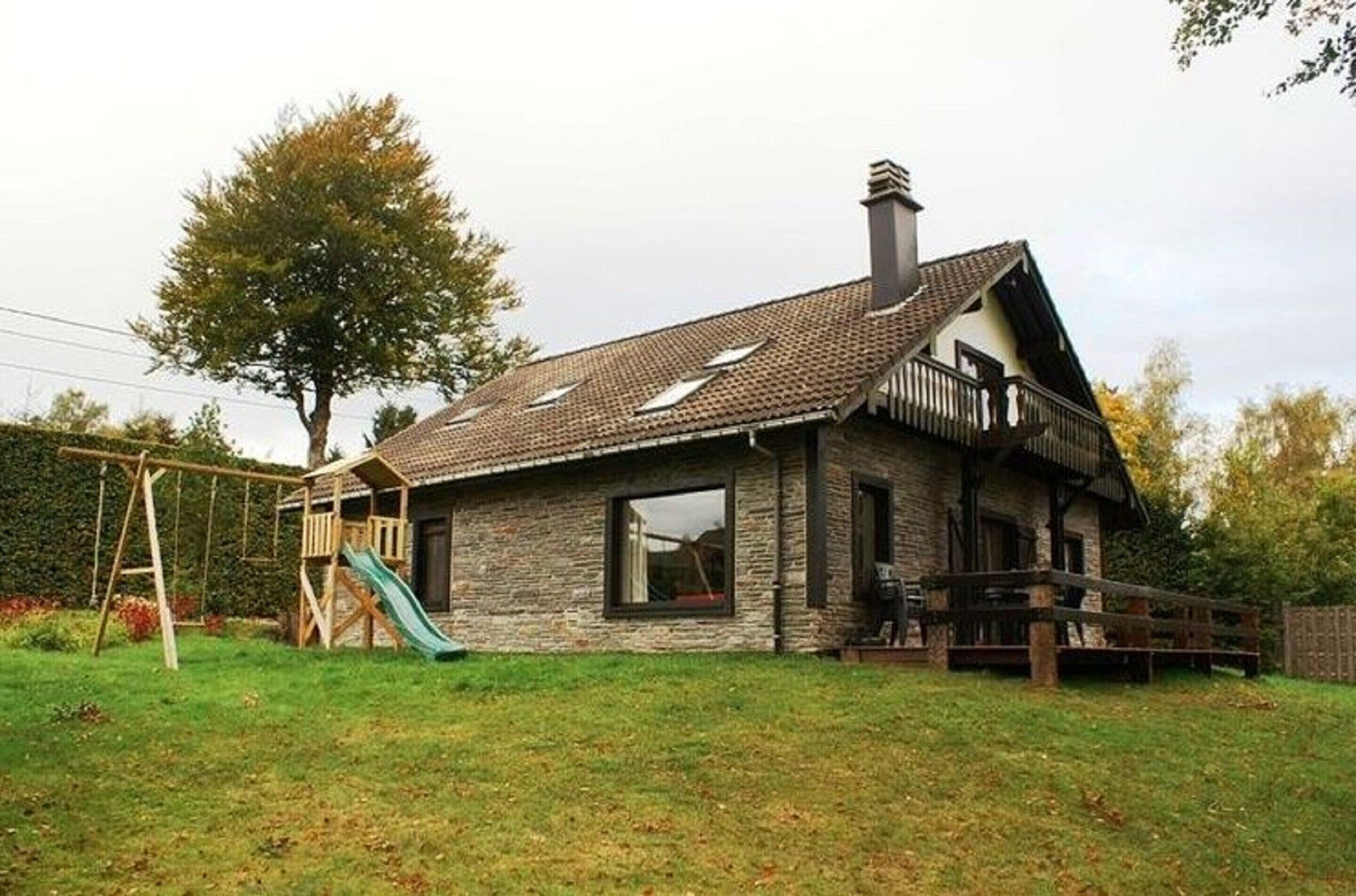 Ein Haus aus Stein mit einem geneigten Dach, einer Holzterrasse und einer Schaukel.