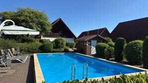 Une piscine avec des chaises longues et un petit bâtiment, le tout entouré d’arbustes et d’arbres bien entretenus.