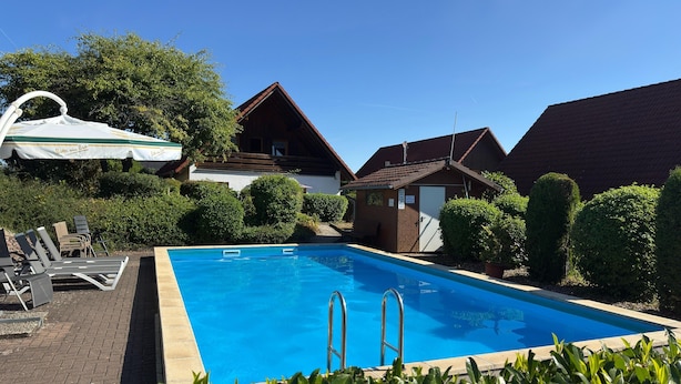 Une piscine avec des chaises longues et un petit bâtiment, le tout entouré d’arbustes et d’arbres bien entretenus.
