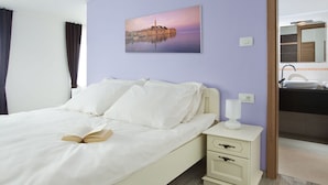 A bedroom with a bed, a bedside table with a lamp, a painting of a cityscape, and a bathroom visible through an open door. - Attractive Villa With Private Swimming Pool, Beach Volleyball and Fenced Garden (Rasa)