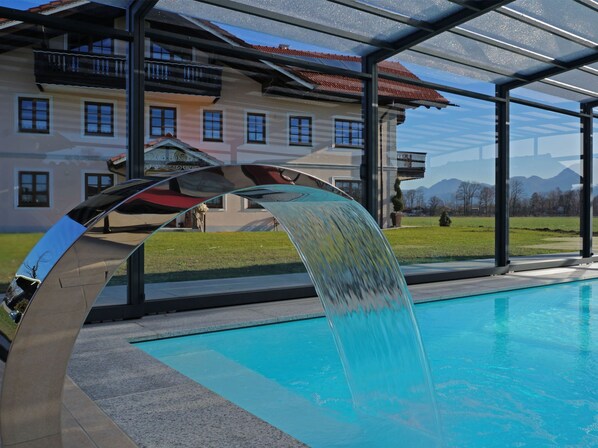 A swimming pool with a modern water feature, a covered area with a glass roof, and a building with a red-tiled roof in the background. - Quaint Holiday Home in Feldwies near Ski Area (Uebersee)