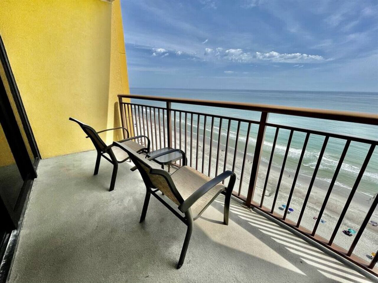 A balcony with two chairs and a table, overlooking a beach with clear skies.