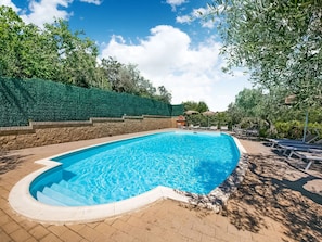 En swimmingpool med en stenvæg og en hæk på den ene side, omgivet af liggestole og parasoller.