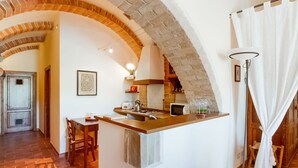Een gezellige keuken met een centraal eiland, zichtbare bakstenen bogen en een eethoek met houten meubels.