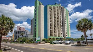 Un hotel alto con fachada verde y color beige, varios pisos y un estacionamiento con automóviles.