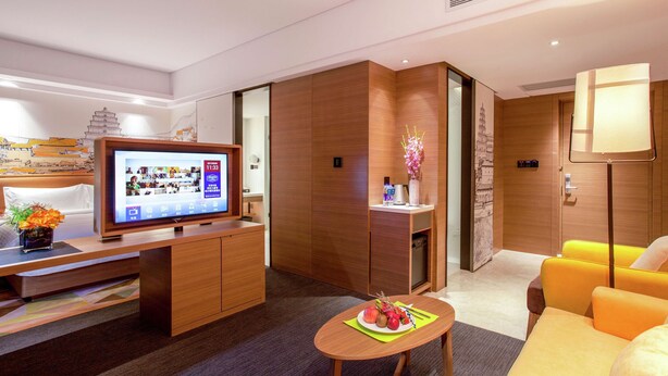 A modern hotel room with a flat-screen TV, wooden furniture, a dining table with fruit, and a sofa. - Hampton BY Hilton Yangzhou Slender (Yangzhou)