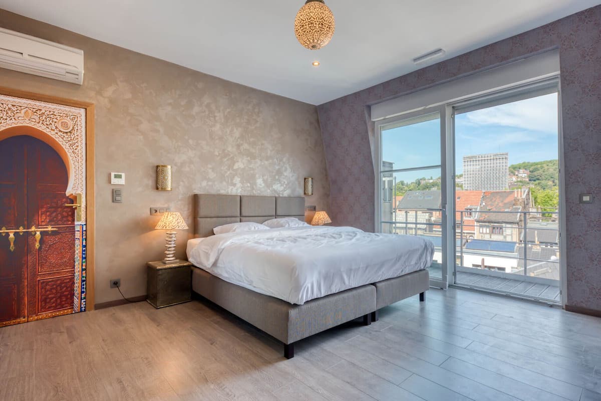 A modern bedroom with a large bed, two bedside lamps, a decorative wall piece, and a view of the city through large windows.