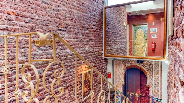 Una escalera con barandillas de metal ornamentadas, un muro de ladrillo y vistas a una habitación con una puerta estampada y decoración tradicional.