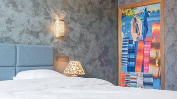 A hotel room with a bed, a wall-mounted lamp, and a colorful painting of buildings.