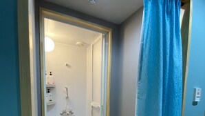 A small, enclosed shower area with a glass door, a blue curtain, and a small shelf with toiletries. - 88HOTELS (Matsuyama)