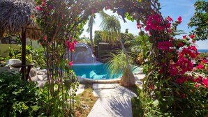 Un espace piscine agrémenté d’une structure à toit de chaume, entouré d’une végétation luxuriante et de fleurs aux couleurs vives.