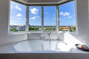 A modern bathroom with a large bathtub, a window with a view of a residential area, and a showerhead.