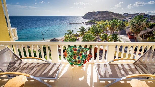 A balcony with lounge chairs overlooking a beach, palm trees, and a mountainous backdrop.
