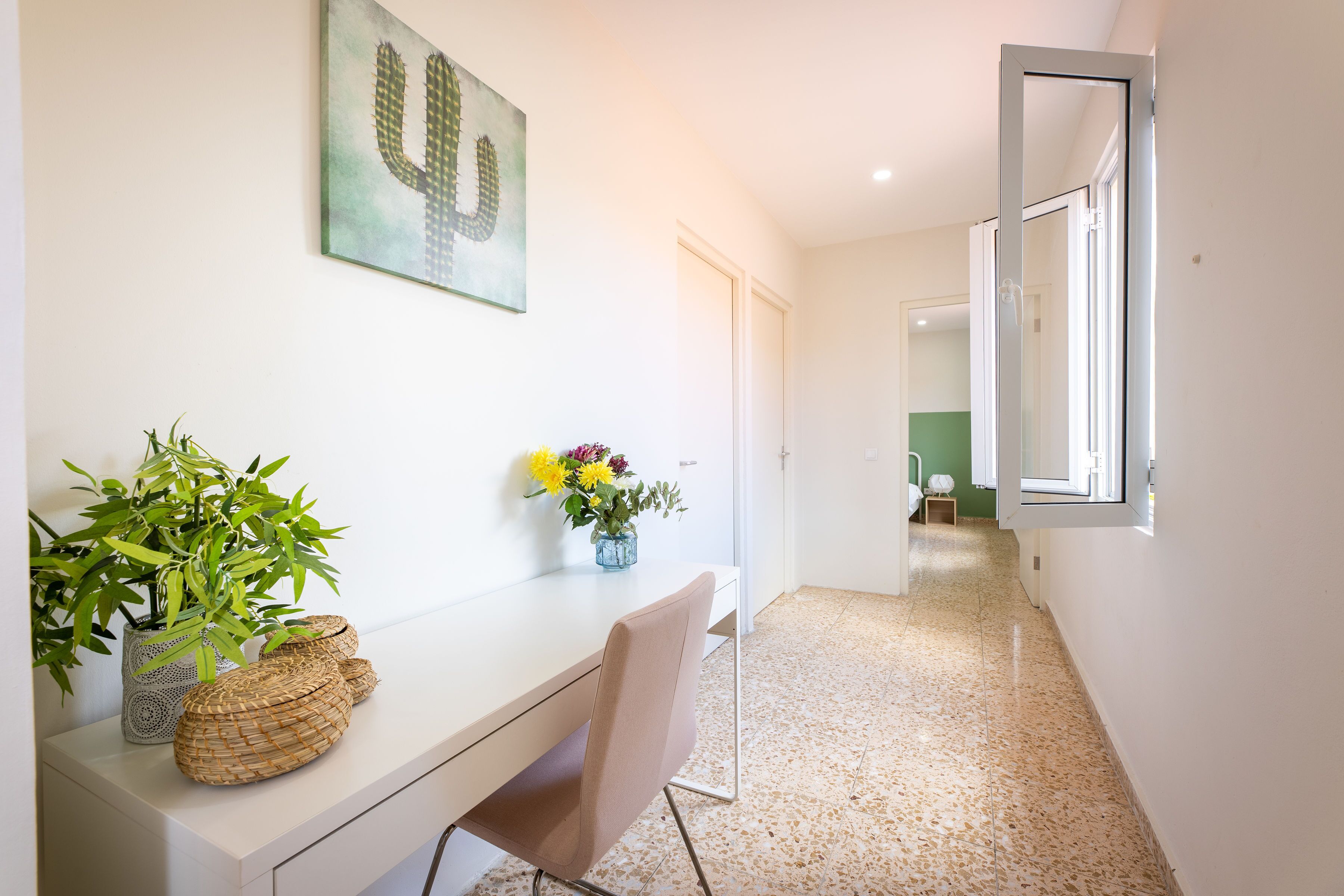 A hallway with a desk, chair, potted plants, and a cactus artwork.