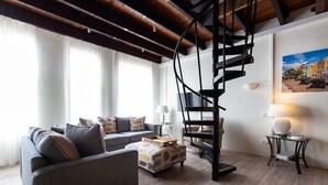 Una sala de estar moderna con un sofá gris, una mesa de centro de madera, una escalera en espiral negra y un cuadro de una escena urbana en la pared.