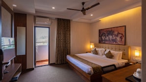 A hotel room with a bed, a desk, a TV, a ceiling fan, and a painting on the wall.