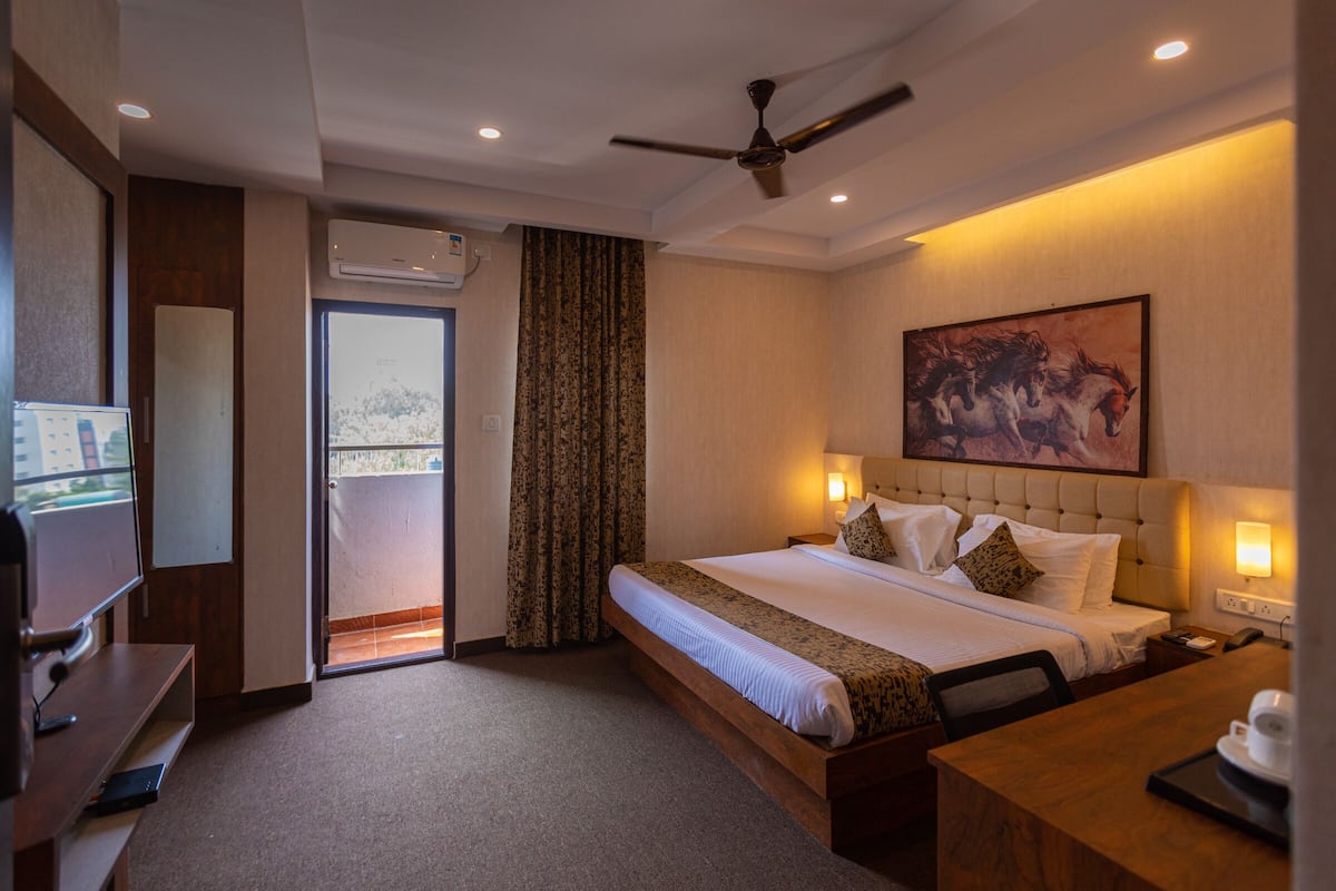 Quarto de hotel com cama, escrivaninha, televisão, ventoinha no teto e um quadro na parede.