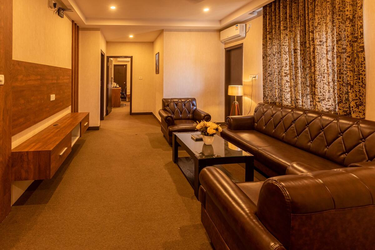 Quarto de hotel com sofá de couro marrom, poltrona combinando, mesa de centro de vidro com um vaso de flores, um banco de madeira e piso carpeteado.